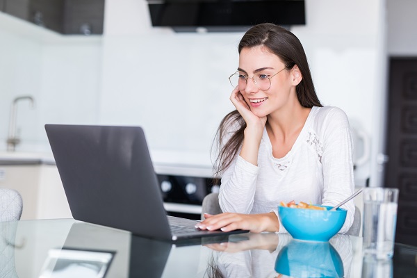 Happy young woman eating salad from a bowl and drinking orange juice while standing on a kitchen and watching movie on laptop